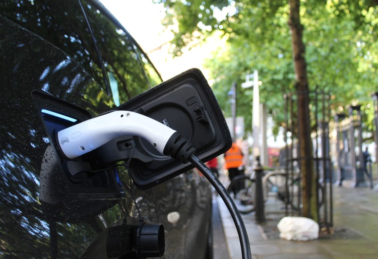 Coche eléctrico recargándose en una estación de carga en una calle arbolada. El conector está enchufado al puerto de carga del vehículo, destacando la infraestructura de carga urbana para vehículos eléctricos. - Carglass.