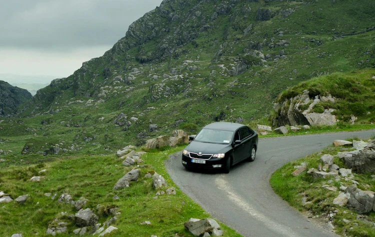 Coche negro conduciendo por una carretera estrecha y sinuosa en una zona montañosa, rodeado de paisajes verdes y rocosos, bajo un cielo nublado.-Carglass