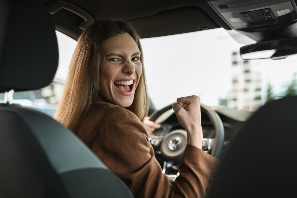 Mujer sonriente celebrando desde el asiento del conductor de su coche nuevo, perfecta para ilustrar consejos para el rodaje de coche nuevo - Carglass.
