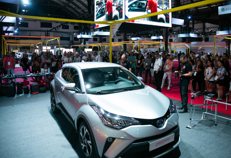 Exhibición de un coche gris en un evento automovilístico con asistentes y pantallas mostrando detalles del vehículo - Carglass.