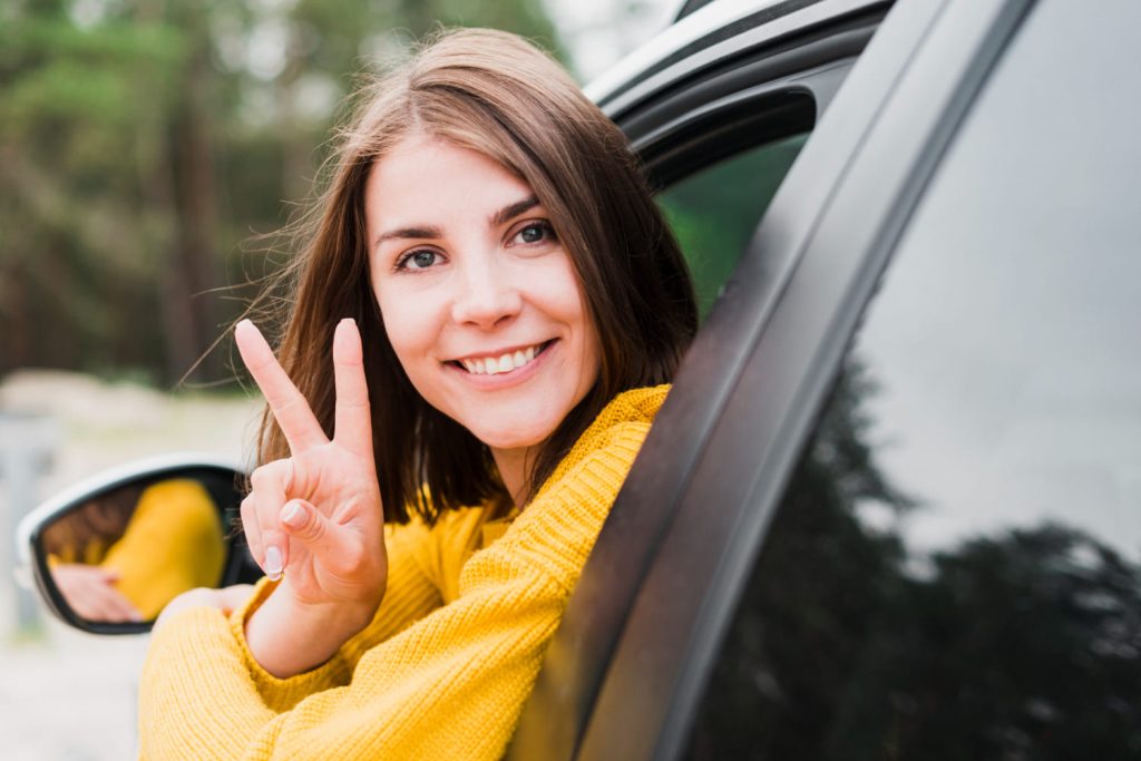 Joven sonriente mostrando un gesto de paz desde la ventanilla de un coche estacionado, ideal para consejos de seguridad vial para conductores noveles - Carglass.