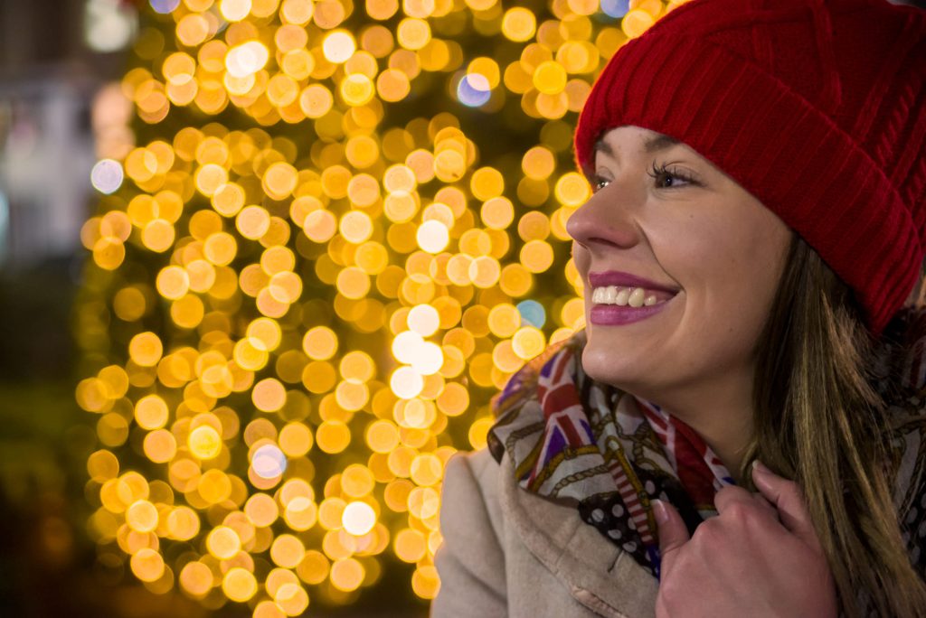 Una mujer sonriente con gorro rojo disfruta de luces navideñas brillantes en rutas de España en diciembre. - Carglass