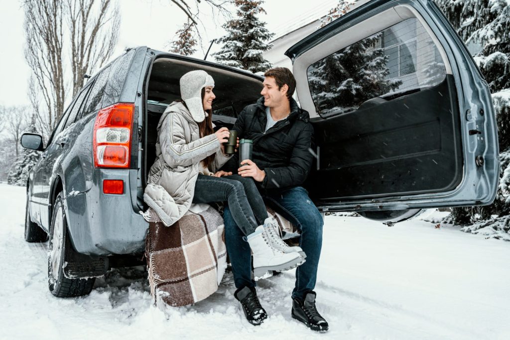 Pareja disfrutando de bebidas calientes junto a su coche en la nieve, resaltando la comodidad y preparación para conducir con nieve en invierno - Carglass.