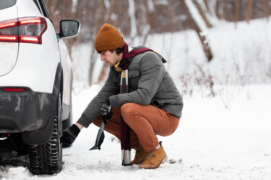 Un hombre revisa la llanta de un coche blanco en un camino nevado, vistiendo ropa de invierno, ilustrando la importancia del mantenimiento coche invierno - Carglass.
