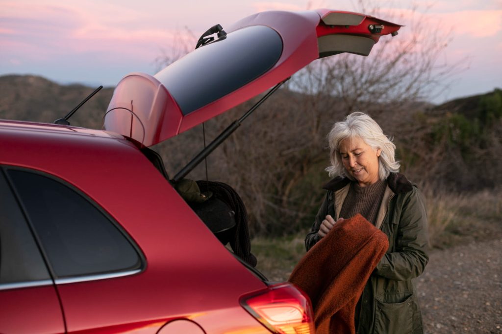 Mujer mayor sacando una manta del maletero de su coche rojo al atardecer. Seguridad en viajes largos y preparación para el frío - Carglass.