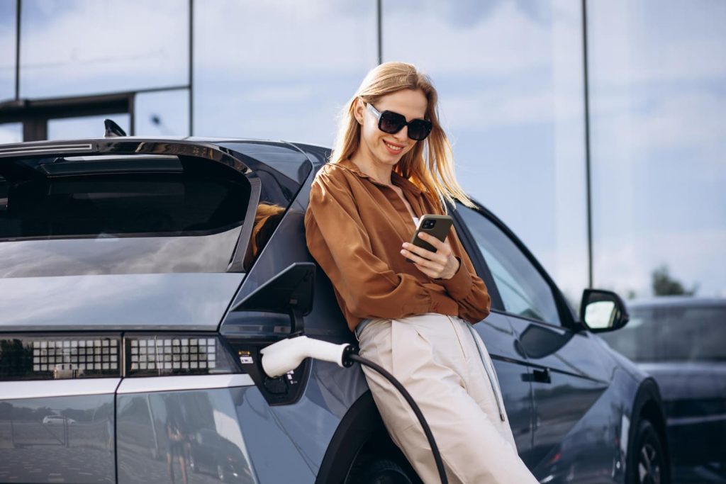 Mujer con gafas de sol sonríe mientras usa su móvil, apoyada en un coche eléctrico en carga. Relacionado con subvenciones Plan MOVES 2025