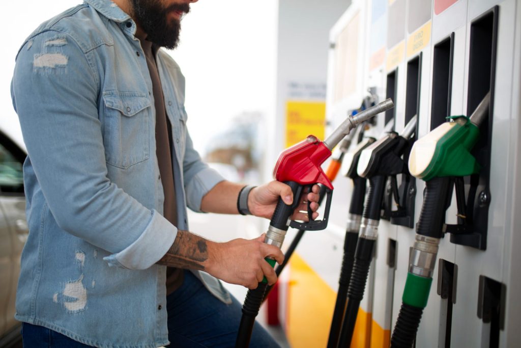 Hombre con camisa de mezclilla recargando gasolina en estación, sostiene boquilla roja para ahorrar combustible - Carglass.